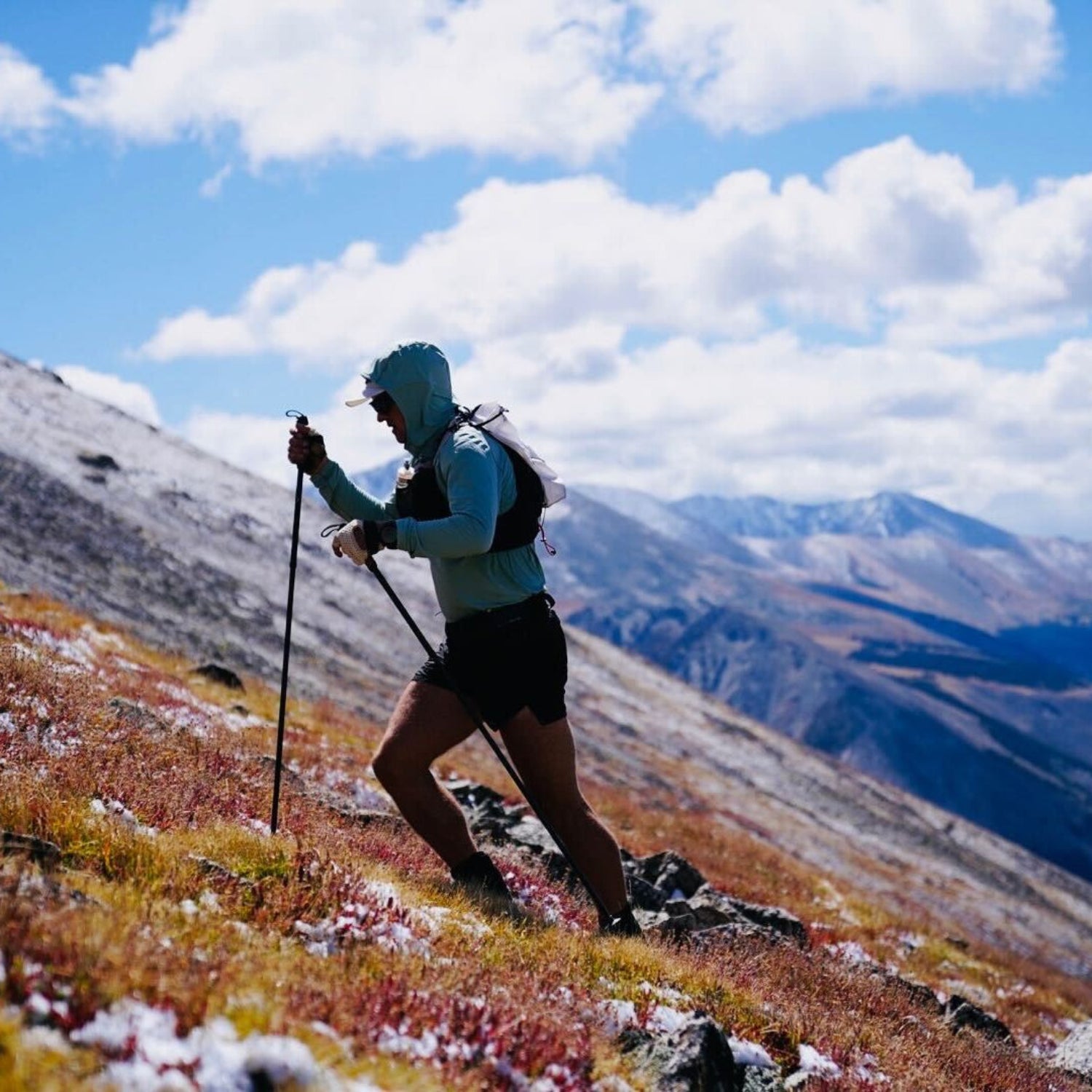 Person hiking on a mountain with backpack and poles, surrounded by snow-capped peaks and blue sky.